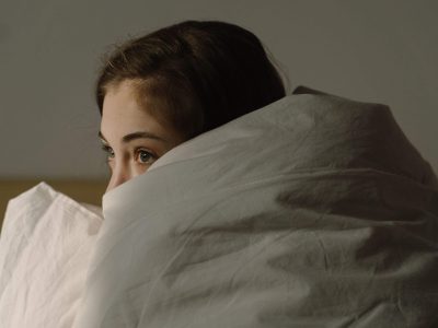 Woman curled up in a blanket with only her eyes visible, representing difficulty fully resting and nervous system activation addressed through sleep therapy for highly sensitive people in Denver, CO.