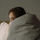 Woman curled up in a blanket with only her eyes visible, representing difficulty fully resting and nervous system activation addressed through sleep therapy for highly sensitive people in Denver, CO.